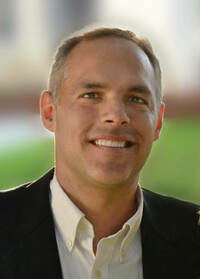 Man wearing white shirt and back coat smiling with blurry grass and building in the background