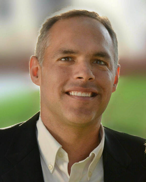 Man wearing white shirt and back coat smiling with blurry grass and building in the background
