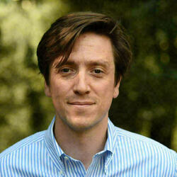 Headshot of a man with medium-length brown hair, wearing a light blue and white striped shirt, slightly smiling against a blurred green, outdoor background.