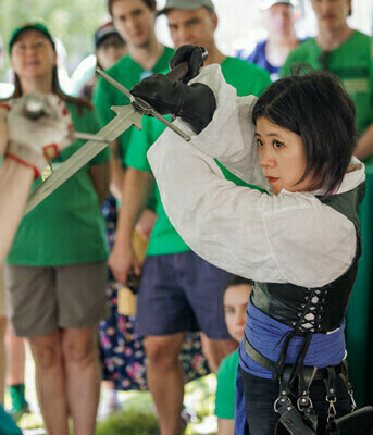 A medieval swordplay demonstrator from Theatrica Gladiatoria wearing a white shirt, black vest, and blue sash holds a sword above their head in a defensive posture. Onlookers in green shirts watch.