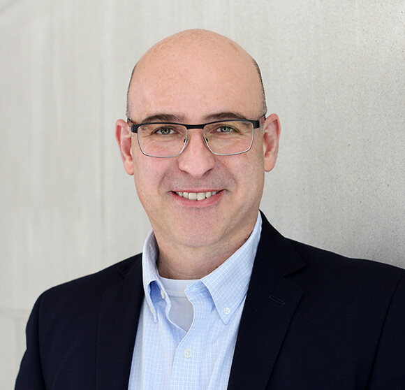 Bald man with glasses wearing a light blue checkered button-down shirt and a dark blue blazer smiles at the camera.