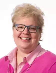 A smiling woman with short, wavy blonde hair and blue-gray framed glasses. She wears a pink collared shirt under a bright pink cardigan, with a thin beaded necklace.