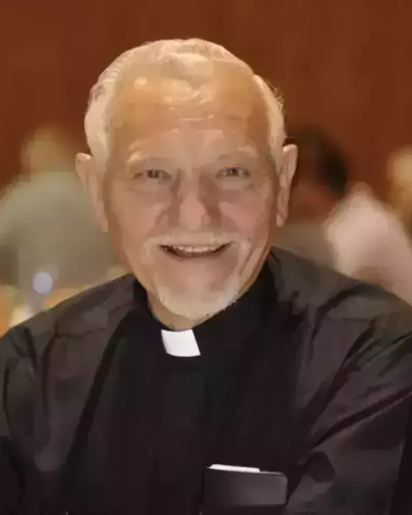 An older man with white hair and a white goatee smiles broadly, wearing a black shirt with a white clerical collar. Blurred background.