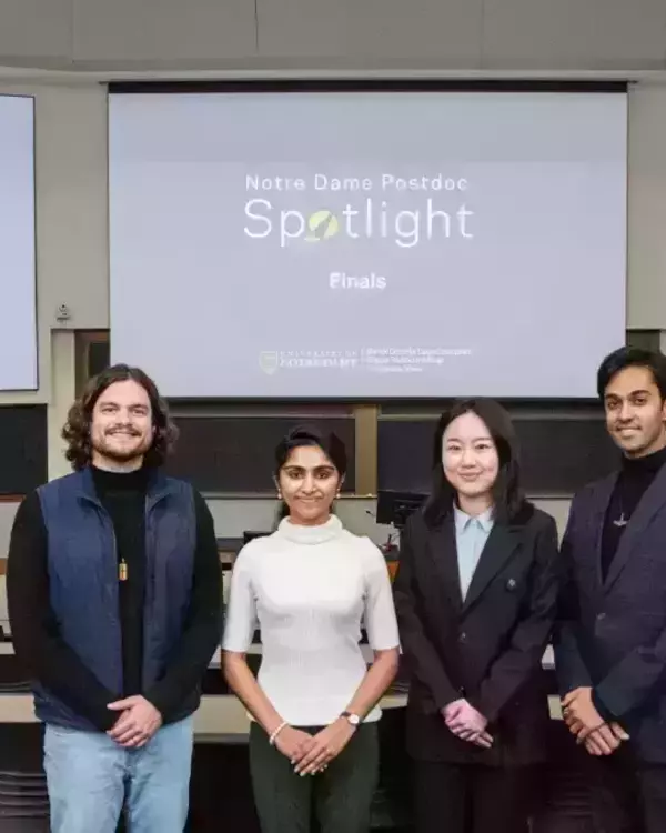 Four smiling finalists for the Notre Dame Postdoc Spotlight stand in a university lecture hall. They pose in front of screens displaying the event title and university logos. Empty classroom desks are visible.