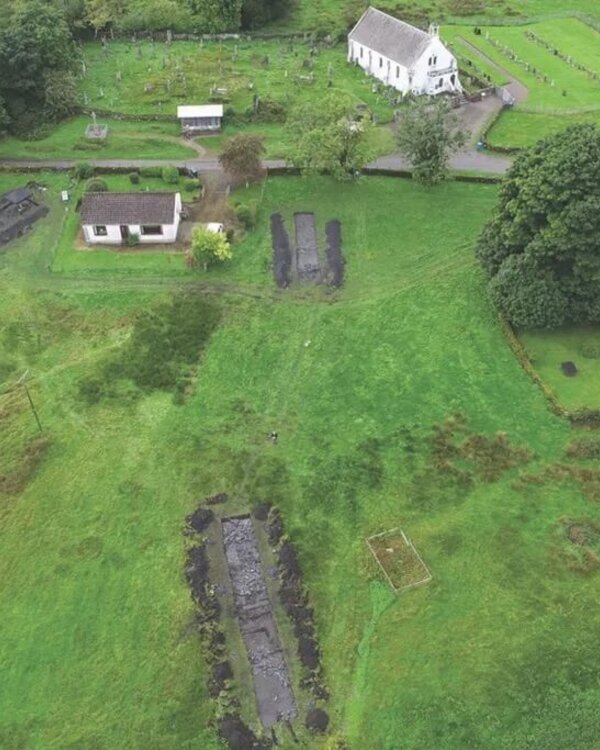 Archaeological excavation sites near a white church and houses in a grassy landscape.